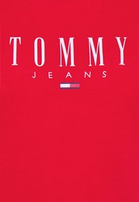 Czerwona koszulka z białym napisem "TOMMY" nad "JEANS". Posiada małą grafikę flagi w kolorach niebieskim, białym i czerwonym. Wykonana z bawełnianego materiału o gładkiej fakturze.