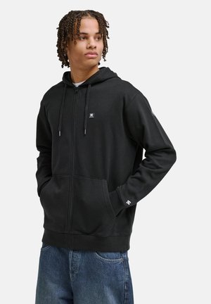 Jonge man met een zwarte hoodie met ritssluiting en handen in de zakken, kijkend naar de zijkant, gecombineerd met blauwe spijkerbroek tegen een effen achtergrond.