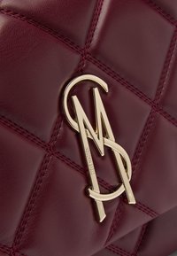 Burgundy kviltet lædertaske med en fremtrædende logoaccent i guldfarve og kontrasterende syninger.