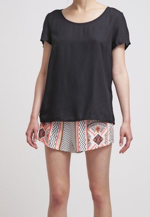 Kvinde iført en løs sort t-shirt med korte ærmer og mønstrede shorts med geometriske designs i orange, sort og hvid.