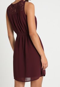 Bordeauxrotes Kleid mit Spitzenbesatz, tailliertem Bund und gefälteltem Rock; aus leichtem Stoff mit glatter Textur gefertigt.