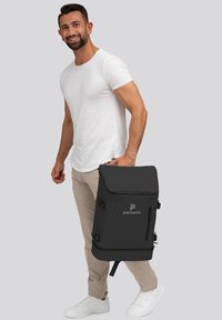 Mochila negra con solapa superior, cremallera lateral y cierres de hebilla, con el logo "pactastic". Material suave y diseño minimalista.