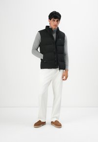 Mand iført sort quiltet vest over grå langærmet trøje, hvid bukser og brune slip-on sko, stående mod en hvid baggrund.