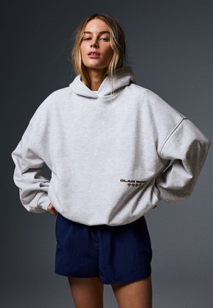 Vrouw die een lichtgrijze oversized hoodie en donkerblauwe korte broek draagt, staat met de handen in de zij tegen een donkere achtergrond.