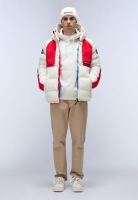 Giacca imbottita bianca e rossa, indossata sopra un hoodie bianco. Pantaloni beige e sneakers bianche con lacci. Berretto lavorato a maglia grigio, design minimale.