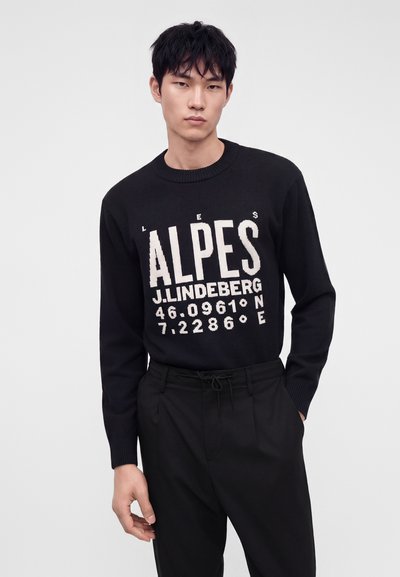 Jeune homme aux cheveux noirs portant un pull noir avec un texte blanc "ALPES J.LINDEBERG 46.0961°N 7.2286°E" et un pantalon noir.