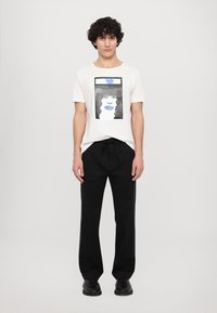 White cotton t-shirt with a graphic print, paired with black trousers featuring a drawstring waist and blue side stripes.