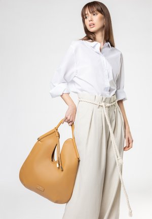 Vrouw draagt wit overhemd met knopen en beige broek met hoge taille houdt een grote bruine leren handtas met koordriem vast tegen een lichte achtergrond.