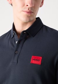 Marineblå polo skjorte laget av bomull, med ribbestrikket krage, tre knapper og et rødt rektangulært merke med "HUGO" i svart broderi.