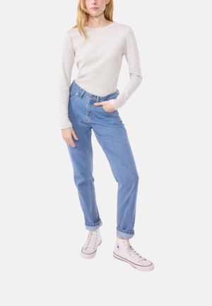 Vrouw staat met één hand in de zak, gekleed in een lichtgrijze lange mouw top, hoge taille blauwe jeans en witte hoge sneakers.