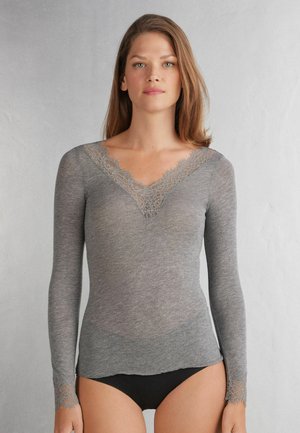 Kvinde iført en tætsiddende langærmet grå top med blondekant ved V-udskæringen og manchetterne, stående mod en ensartet lys baggrund.