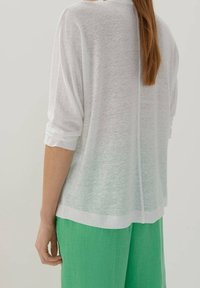 Vit, halvgenomskinlig långärmad topp med en avslappnad passform och uppvikta ärmar. Stylen parats med gröna, lätta, vida byxor.