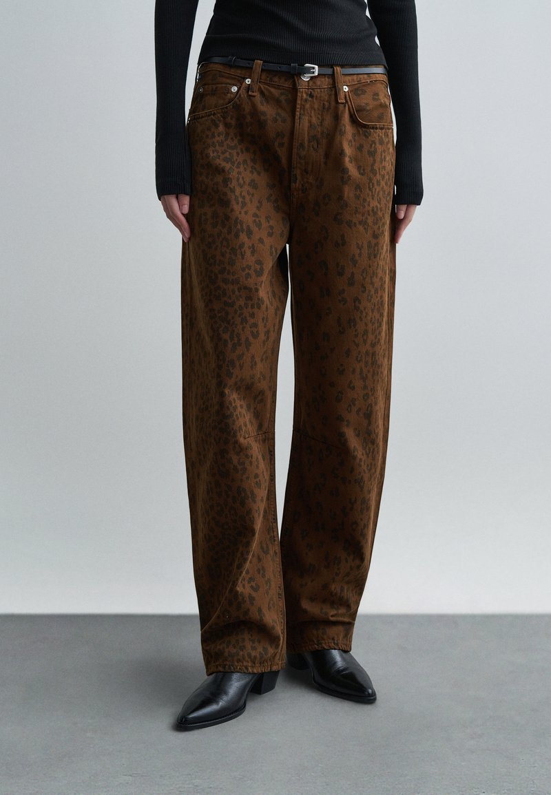 Calças largas com estampa de leopardo castanha, bolsos laterais, detalhes costurados e cinto. Combinadas com botas pretas de bico fino até ao tornozelo.