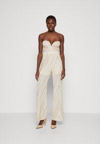 Beige, trägerloser Jumpsuit mit V-Ausschnitt und weitem Bein, versehen mit metallisch goldenen Zickzackmustern und taillierter Passform. Nackte High Heels runden den Look ab.