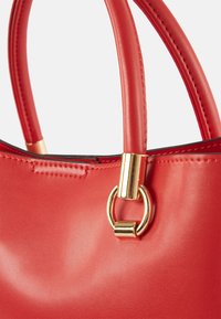 Sac à main en cuir rouge avec poignées rondes et détail anneau métallique doré reliant la poignée au corps du sac.