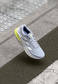 Baskets de course grises claires avec un upper texturé, des rayures blanches et une semelle extérieure jaune vif. Présente un système de laçage et un col rembourré.