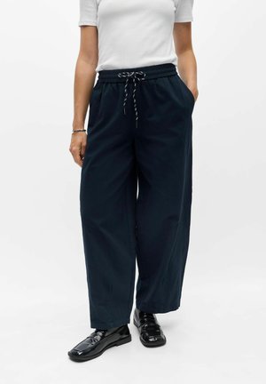Person iført vidde, navy bukser med snøreliv, hvid kortærmet top, sorte skinnende loafers og den ene hånd i lommen.