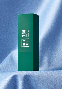 Grønn rektangulær leppestiftkasse med matt finish, som har hvit tekst: "734 LEPPESTIFT" og logo på den ene siden. Myk blå teksturert bakgrunn.
