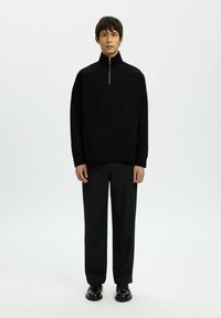 Pull noir avec col montant et fermeture éclair. Porté avec un pantalon noir ajusté et des chaussures noires, présentant une texture lisse et un design minimaliste.