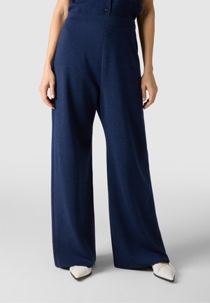 Kobieta w granatowych spodniach o szerokich nogawkach i białych butach w szpic, stojąca z rękami swobodnie opuszczonymi wzdłuż ciała.