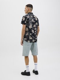 Chemise noire à manches courtes avec motif floral blanc, associée à un short en denim bleu clair. Modèle portant des chaussures noires et des chaussettes blanches.