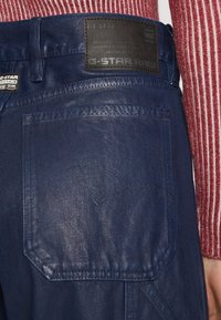 Dark blue denim jeans with a textured finish, featuring a back pocket and a G-Star Raw brand label at the waistband.