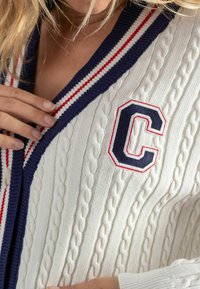Witte kabelgebreide cardigan met marineblauwe en rode gestreepte afwerking, met een geborduurd marineblauw letter "C" op de linkerkant. Gevoelige stoffen details zijn zichtbaar.