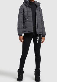 Doudoune grise avec capuche, poignets côtelés, fermeture éclair et deux poches avant. Portée sur un col roulé noir et associée à des leggings et des bottes noirs.