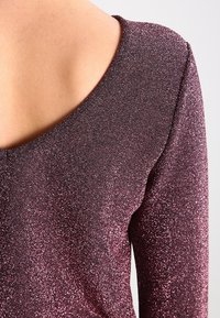 Top in velluto viola lucido con una profonda scollatura sul retro, dotato di maniche lunghe e una texture che riflette la luce, evidenziando la sua qualità scintillante.