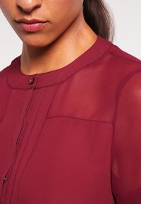 Blouse bordeaux avec des manches transparentes, encolure ronde et détail de bouton à l'avant. Présente des plis verticaux et une texture lisse.