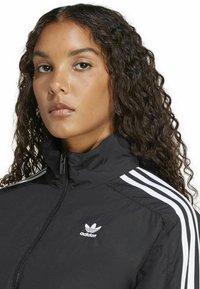 Jachetă neagră cu fermoar, cu guler înalt, având trei dungi albe pe fiecare umăr și un mic logo Adidas alb pe piept.
