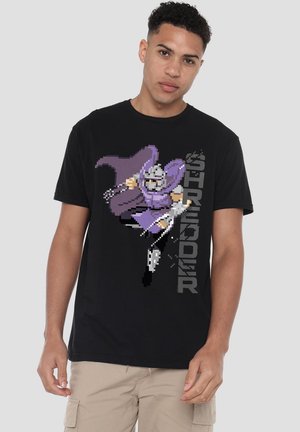 Young man wearing black t-shirt with pixel art of a purple-cloaked warrior and vertical "Shredder" text, paired with beige cargo shorts.