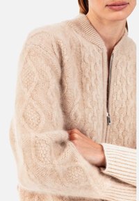 Cardigan beige en maille torsadée avec fermeture éclair à l'avant, fabriqué dans un matériau doux. Il présente des poignets côtelés et un col rond, mettant en valeur des motifs texturés.