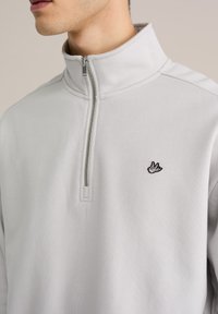 Bluză cu fermoar scurt, de culoare gri deschis, cu textură moale, având un logo brodat cu pasăre pe pieptul stâng. Închidere cu fermoar la gât.