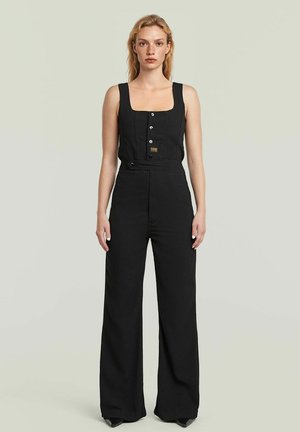 Frau mit hellem Haar, die einen ärmellosen schwarzen Jumpsuit mit Knöpfen und weitem Bein trägt, steht vor einem schlichten hellen Hintergrund.