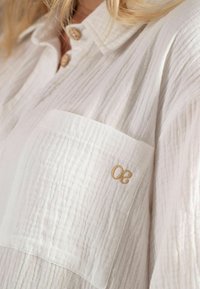 Witte button-up shirt van een gestructureerde, gekreukte stof. Heeft een borstzak en een gouden geborduurd detail "30" op de zak.