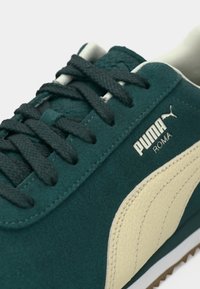 Πράσινη σουέτ Puma Roma αθλητικά παπούτσια με μπεζ δερμάτινες λεπτομέρειες, μαύρες επίπεδες κορδόνες και καουτσούκ σόλα. Λογότυπο ορατό στην εξωτερική πλευρά.