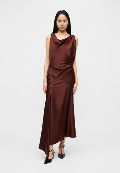 Φόρεμα χωρίς μανίκια από σατέν σε βαθύ μπορντό χρώμα με ντραπέ λαιμόκοψη, ασύμμετρο τελείωμα και πιετά σχέδια· συνδυασμένο με μαύρα παπούτσια με τακούνι.