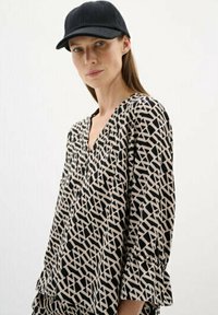 Black and beige patterned blouse with a V-neck and three-quarter sleeves, paired with a black cap. Textured fabric with geometric design.