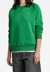 Sudadera verde con cuello redondo y puños acanalados. La sudadera presenta un pequeño logo en el pecho y un ajuste relajado.