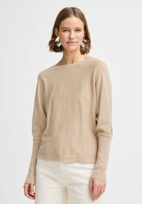 Pull léger beige avec un col large et des manches longues. Texture douce avec une coupe décontractée, associé à un pantalon blanc cassé.