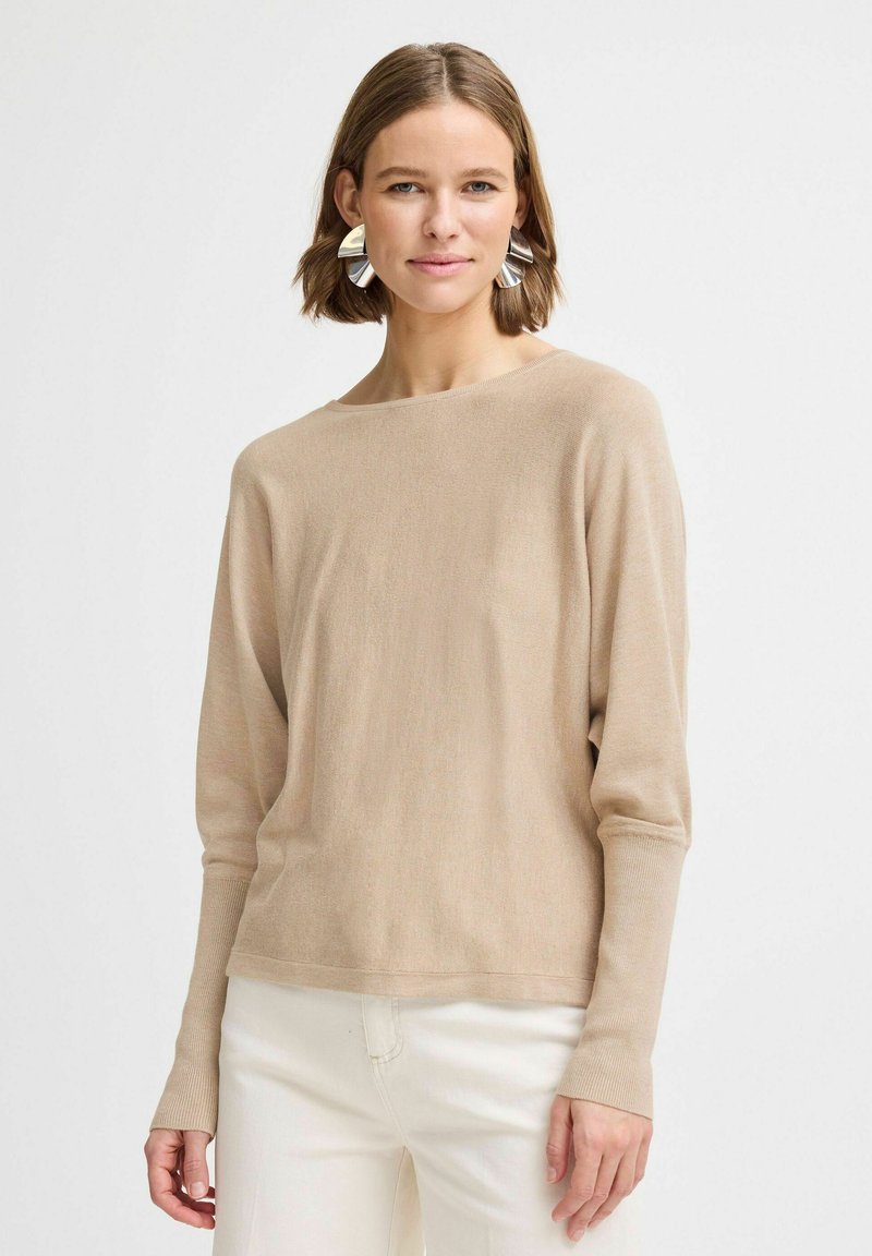Pull léger beige avec un col large et des manches longues. Texture douce avec une coupe décontractée, associé à un pantalon blanc cassé.