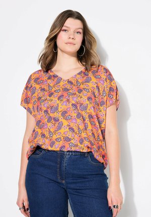 Blusa a maniche corte con scollo a v di colore arancione con un motivo floreale e paisley. Tessuto morbido, vestibilità ampia, abbinata a jeans a vita alta di colore blu scuro.