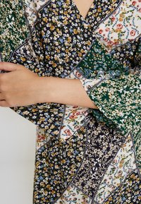 Vestido con estampado floral que presenta una mezcla de telas en verde, azul marino y blanco. Texturizado con pequeñas flores y acentos geométricos. Mangas amplias.