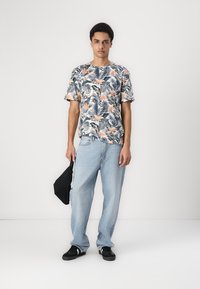 Cămașă cu mânecă scurtă, cu imprimeu tropical, având frunze verzi și flori roz, asociată cu blugi din denim albastru deschis și adidași negri.