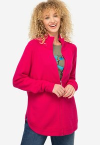 Cardigan fucsia con zip, colletto alto, polsini a costine e texture morbida. Indossato sopra una t-shirt grafica verde e abbinato a jeans.
