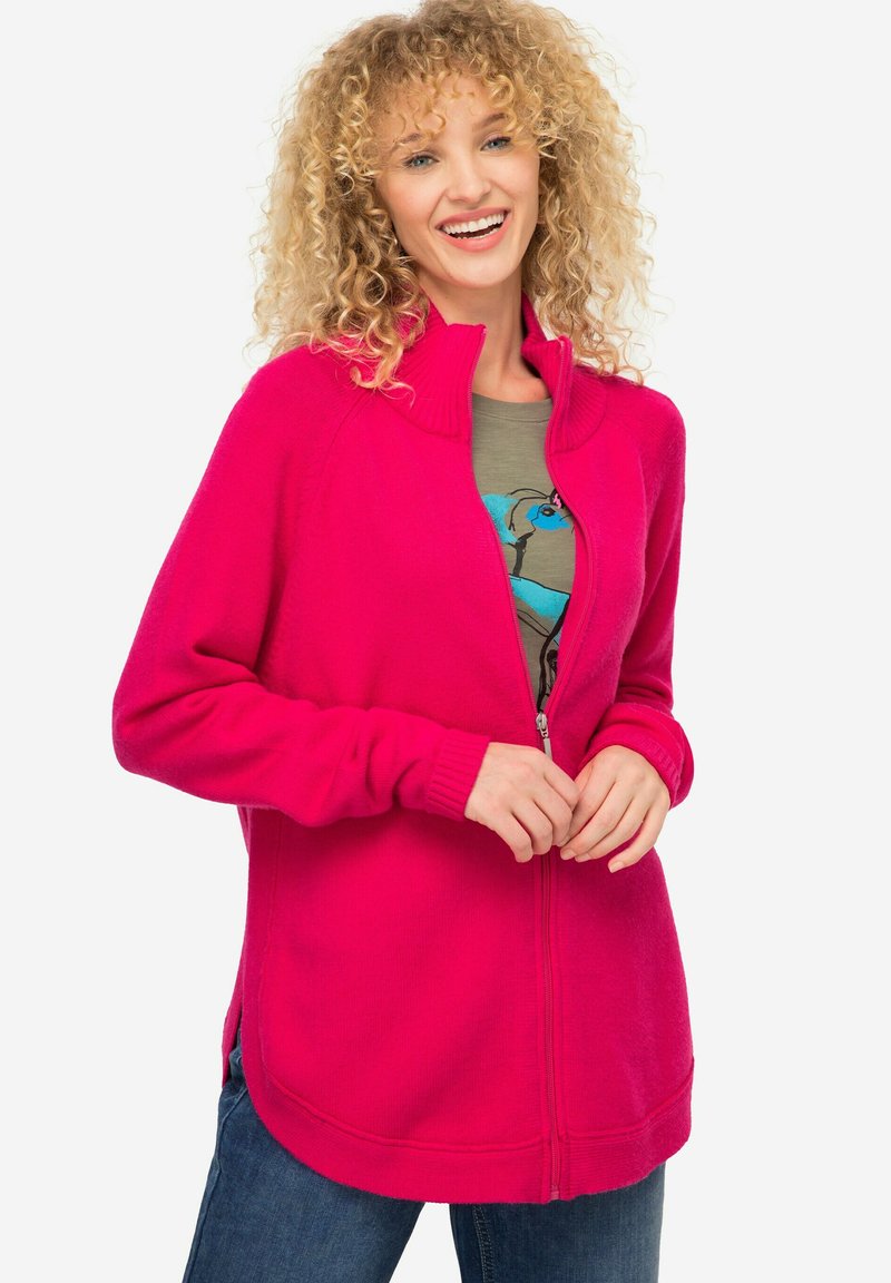 Cardigan fucsia con zip, colletto alto, polsini a costine e texture morbida. Indossato sopra una t-shirt grafica verde e abbinato a jeans.