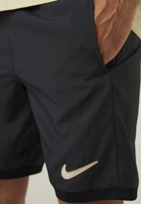 Shorts Nike en tissu noir, avec un logo swoosh brodé en or, des poches latérales et une ceinture noire texturée.