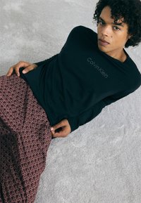 Melna garo piedurkņu krekls ar "Calvin Klein" logotipu, kombinēts ar bordo rakstainām biksēm, kurās redzami ģeometriski motīvi un tekstūra.