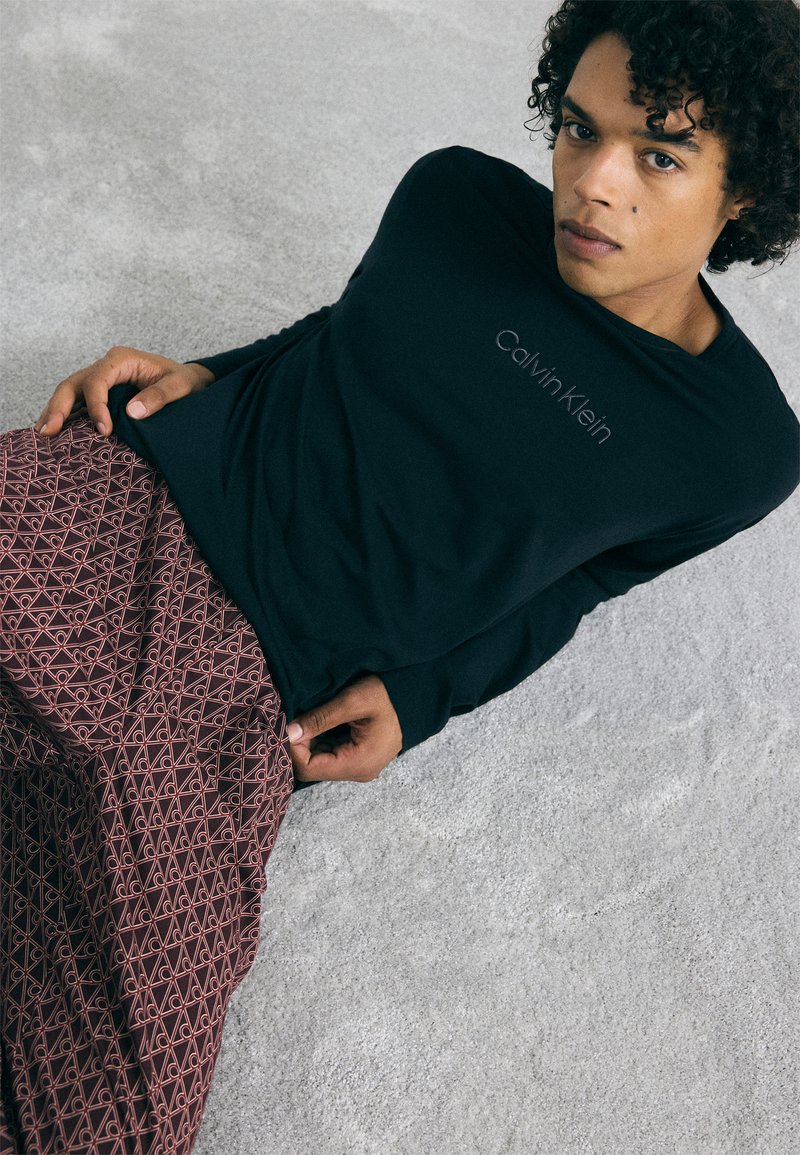 Melna garo piedurkņu krekls ar "Calvin Klein" logotipu, kombinēts ar bordo rakstainām biksēm, kurās redzami ģeometriski motīvi un tekstūra.